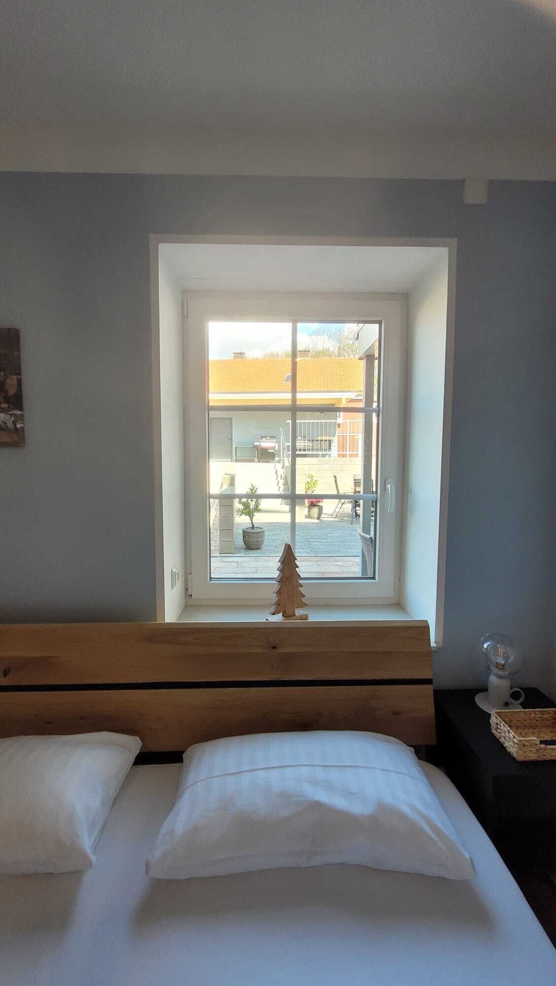 Et soveværelse med træsenggavl, seng med hvidt sengetøj, et lille juletræ i træ samt et vindue med udsigt til en gårdsplads med udendørs møbler og planter.