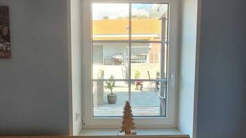 Et soveværelse med træsenggavl, seng med hvidt sengetøj, et lille juletræ i træ samt et vindue med udsigt til en gårdsplads med udendørs møbler og planter.