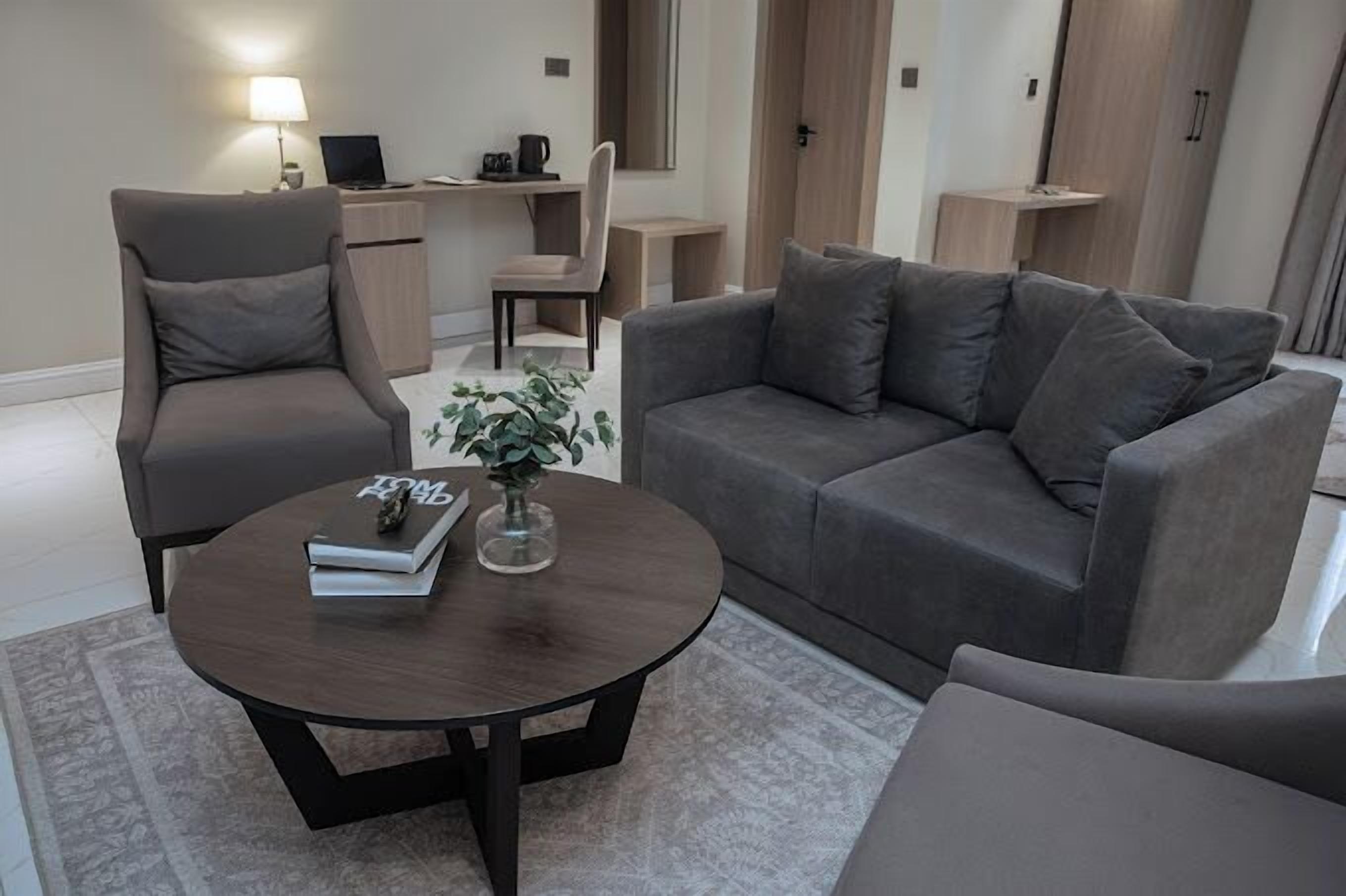 Ein modernes Wohnzimmer mit einem grauen Sofa, einem passenden Sessel, einem runden Couchtisch und einem kleinen Beistelltisch mit Lampe.