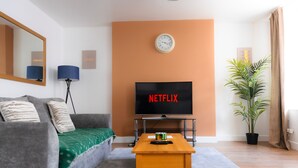 A living room with a sofa, a coffee table, a TV displaying 'NETFLIX', a clock, a lamp, and a plant.