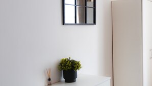 A white wall with a black-framed mirror and a small potted plant with yellow flowers.