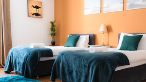 A hotel room with two beds, each with a blue blanket, white pillows, and a green pillow. There is a plant on a side table, a lamp, and artwork on the wall.