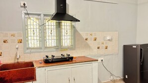 A compact kitchen with a gas stove, a black range hood, a refrigerator, and a window with decorative tiles. - Cozy Beachside Stay Near Mahabalipuram Monuments. (Mahabalipuram)
