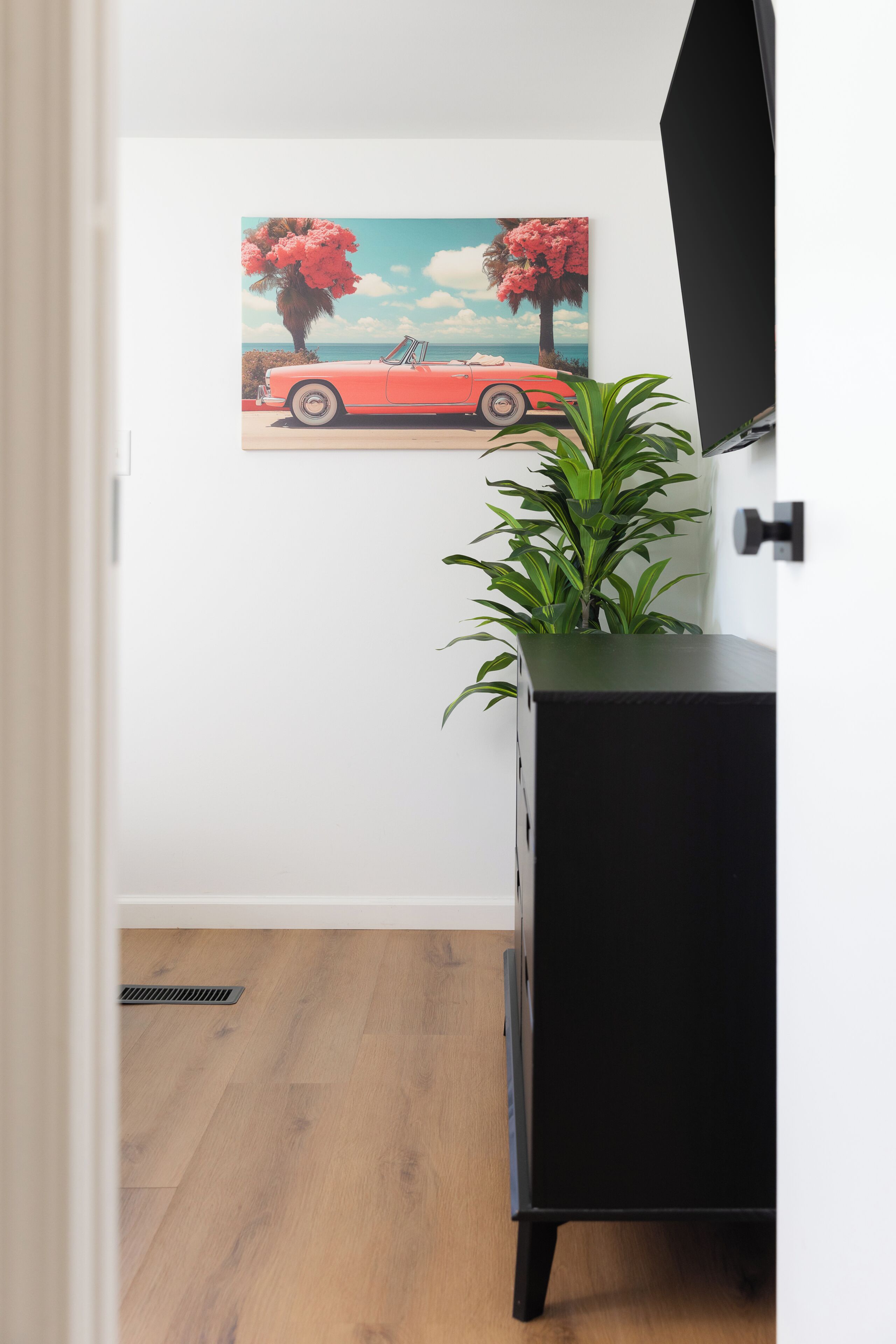 A room with a black cabinet, a potted plant, and a painting of a red car on the wall.