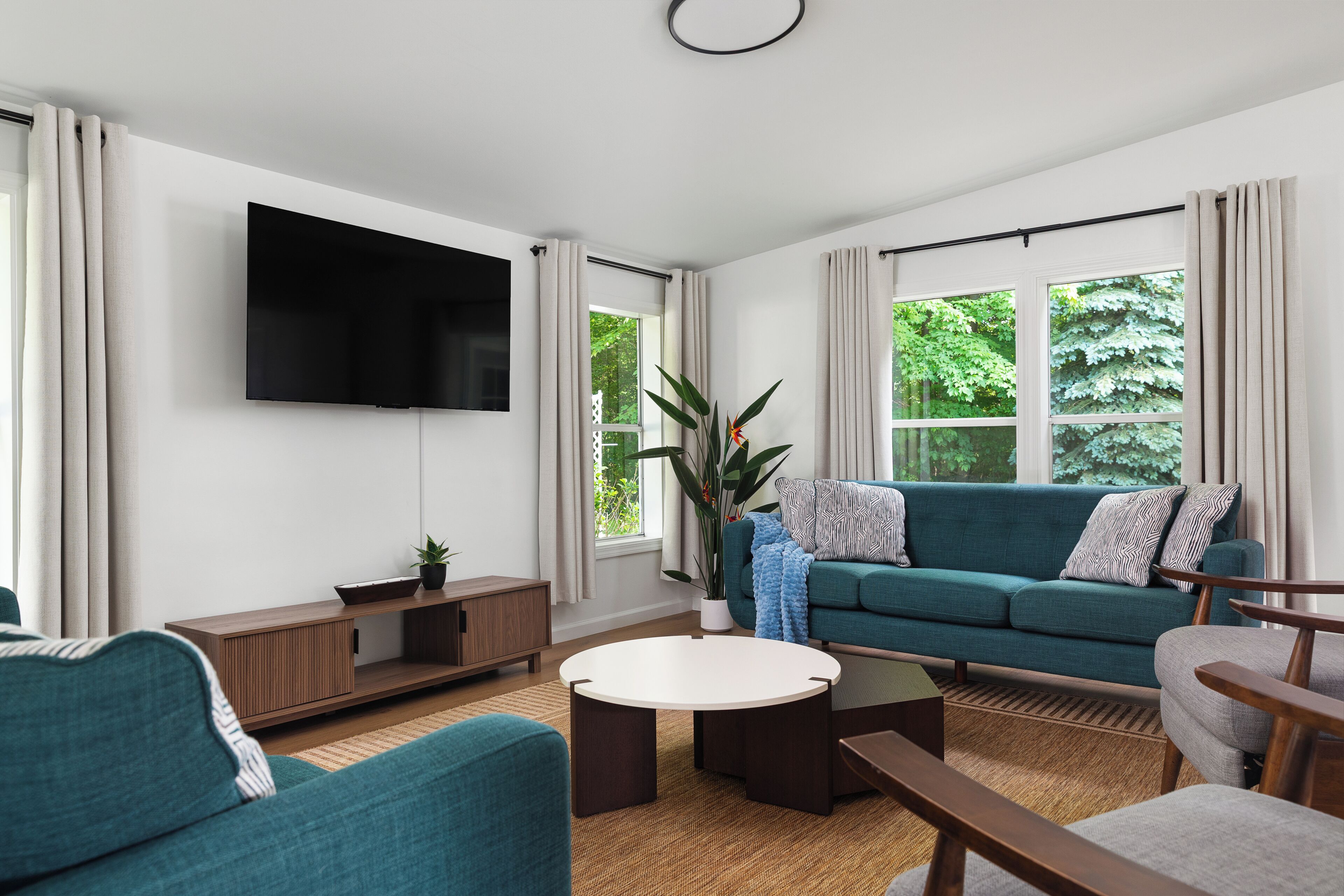 A modern living room with a teal sofa, a flat-screen TV, a coffee table, and a large window with curtains.