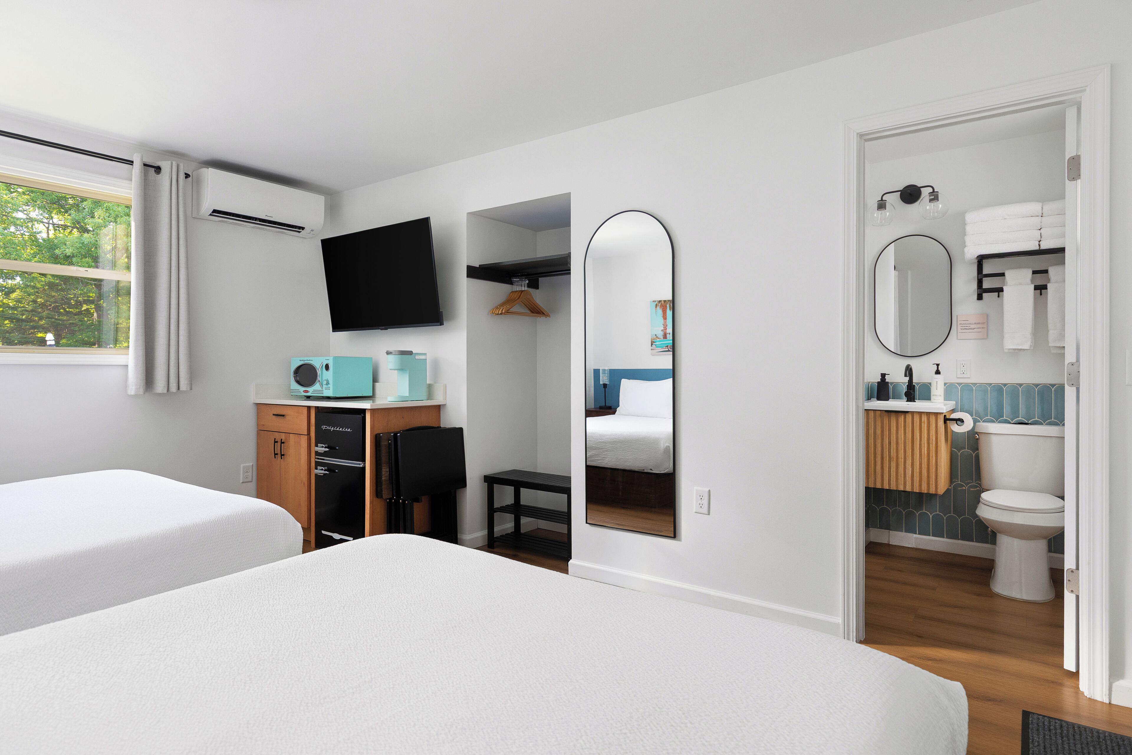 A hotel room with two beds, a TV, a desk with a chair, a mirror, and a bathroom with a toilet and sink.