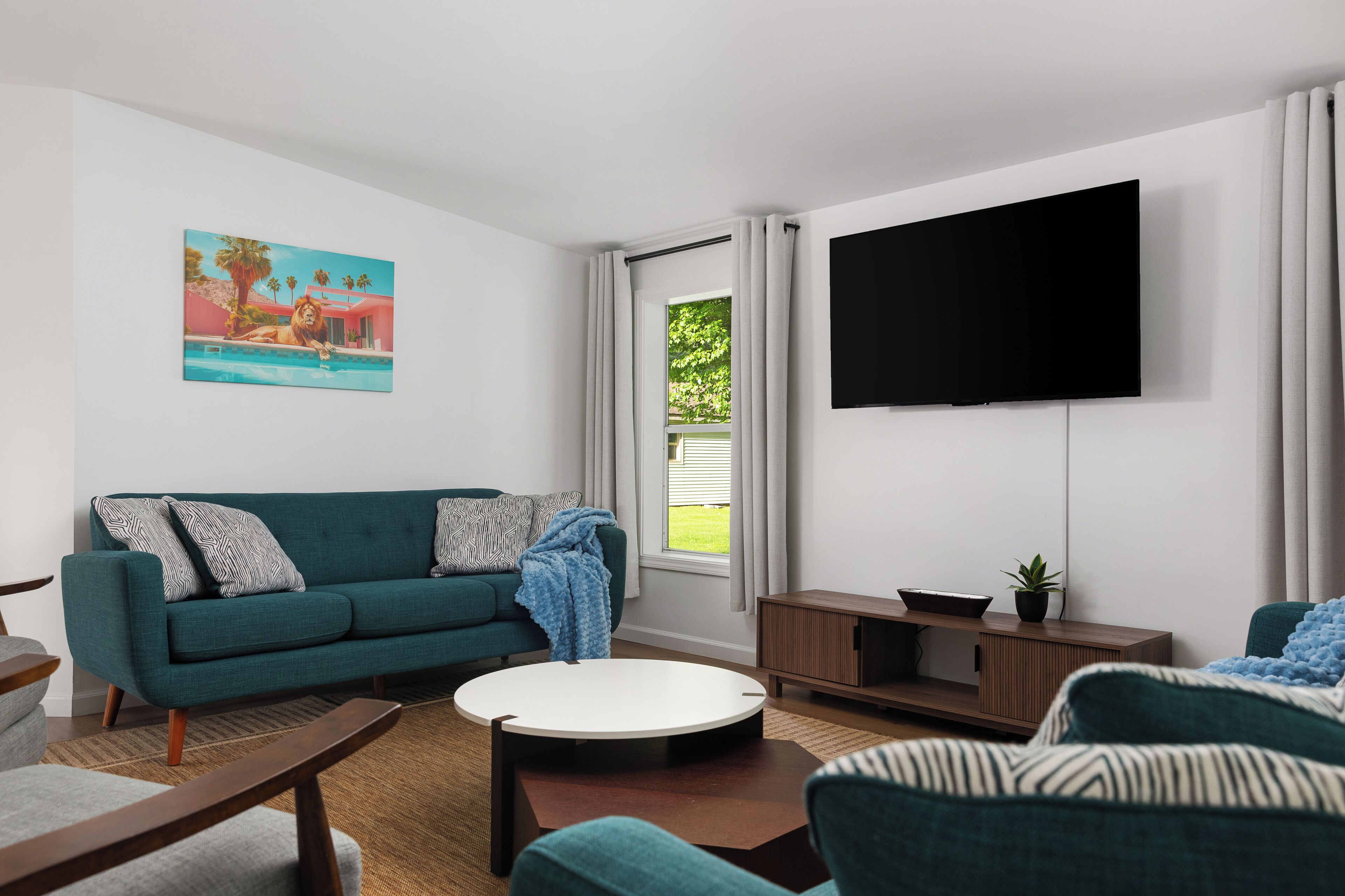 A living room with a teal sofa, a round coffee table, a flat-screen TV, and a painting of a lion.