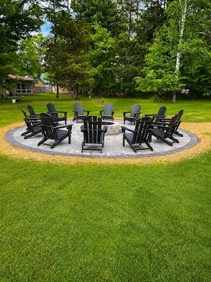 Un espacio circular al aire libre con sillas Adirondack y un brasero central, rodeado de césped cuidado y árboles.