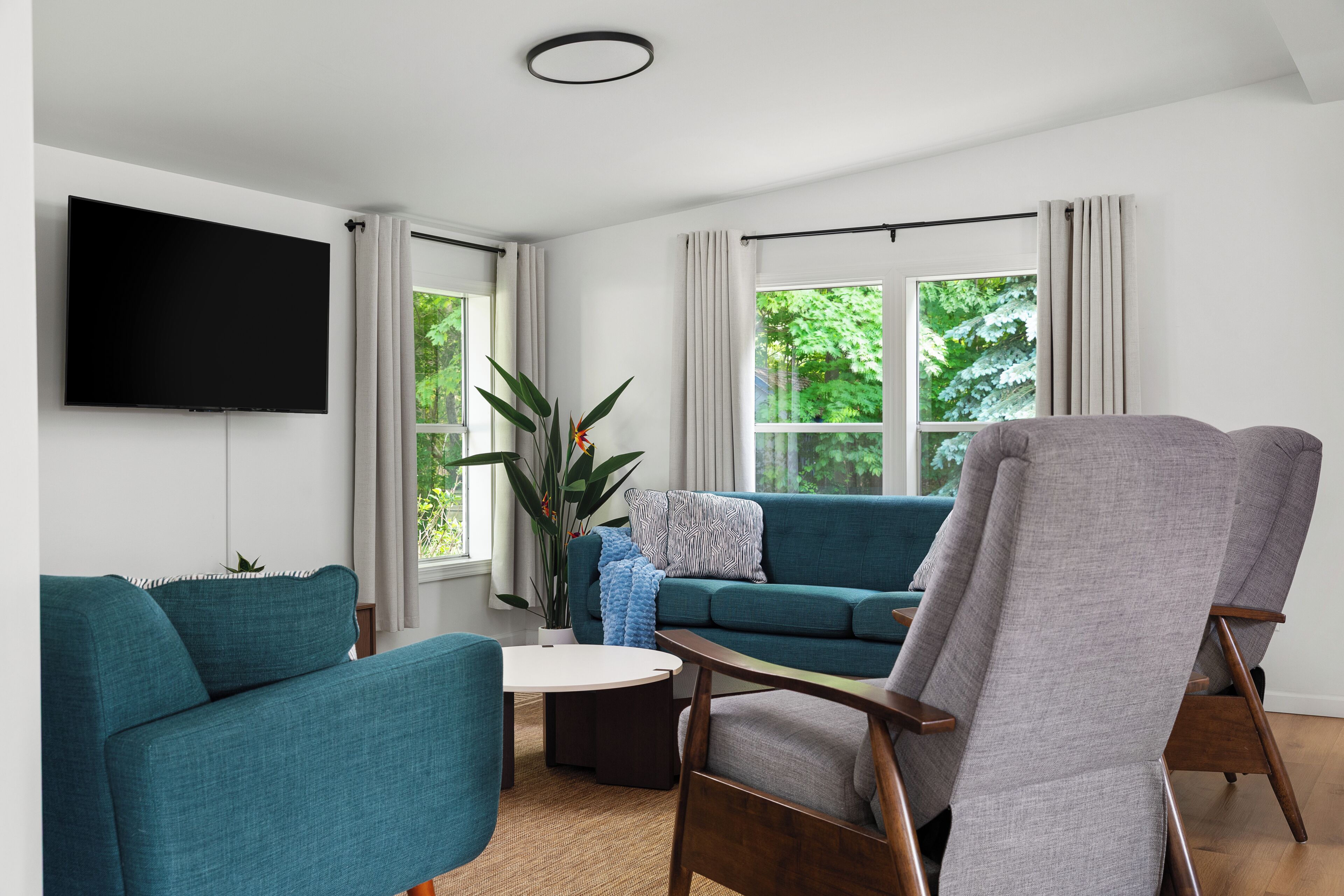 A modern living room with a teal sofa, a grey armchair, a round coffee table, and a flat-screen TV mounted on the wall.