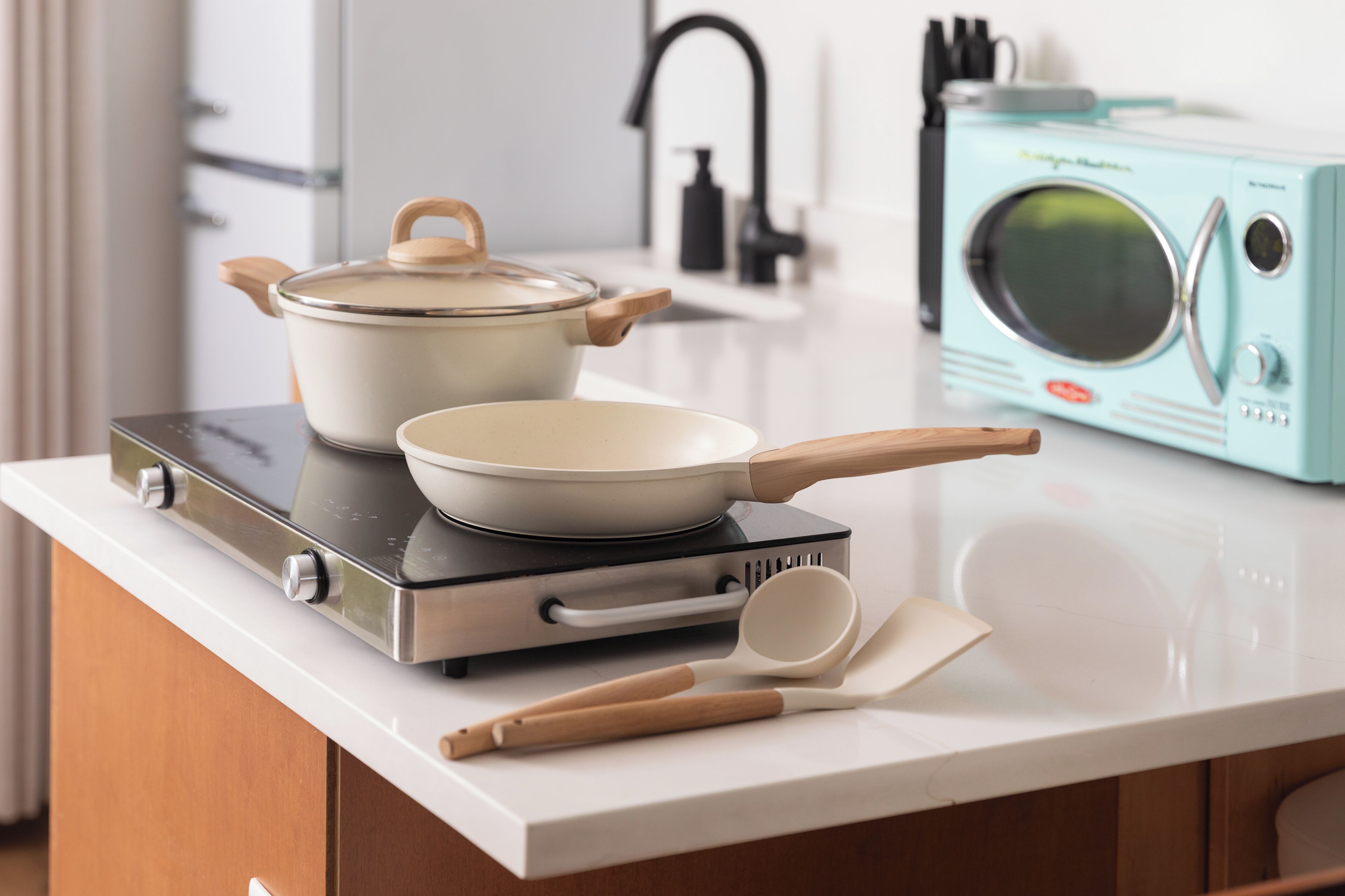 A kitchen with a stove, a pot, a pan, a microwave, and wooden utensils on a countertop.