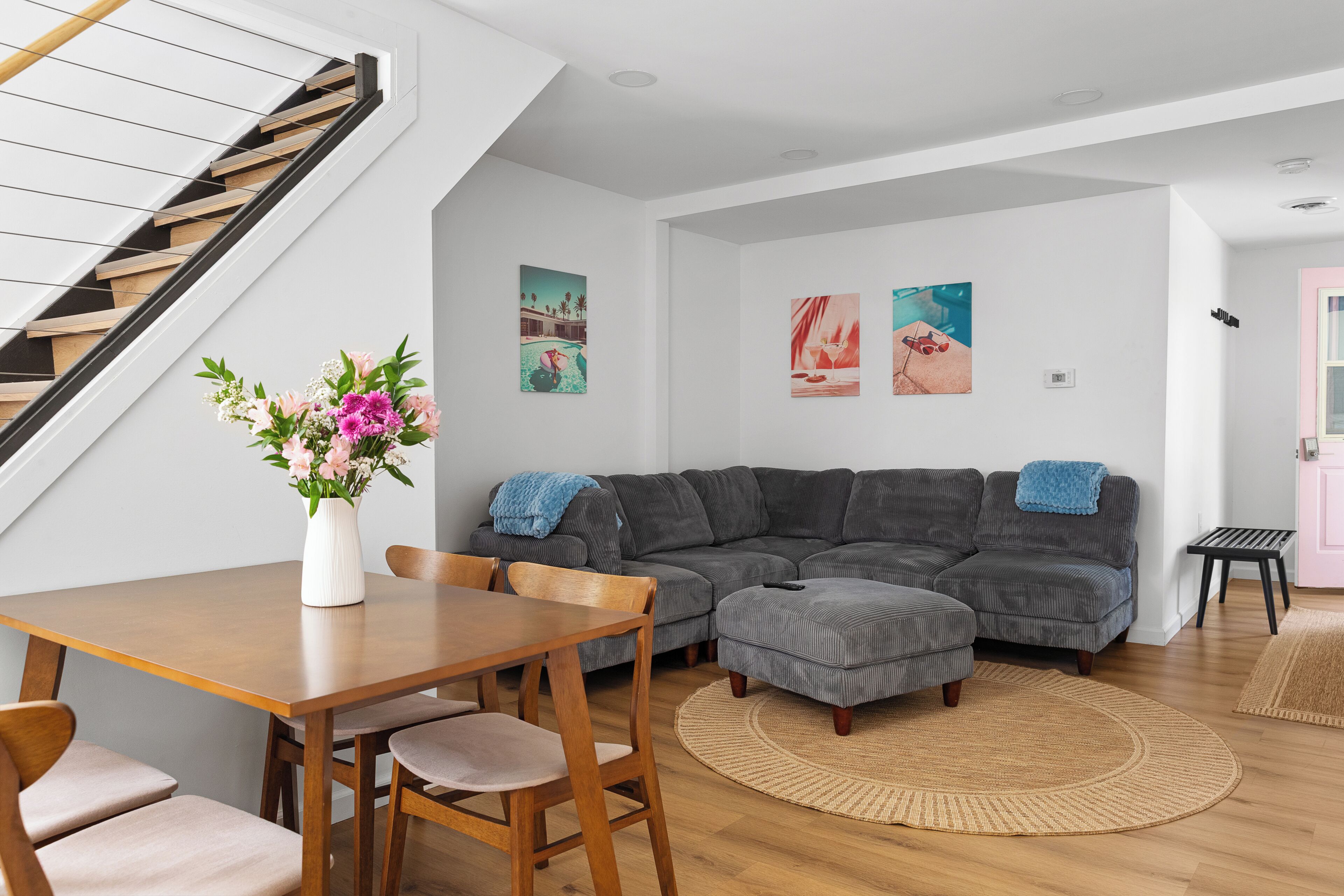 Een moderne woonkamer met een trap, een grijze modulaire bank, een houten eettafel met stoelen, en kunstwerken aan de wanden.