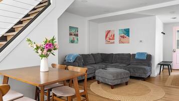 Een moderne woonkamer met een trap, een grijze modulaire bank, een houten eettafel met stoelen, en kunstwerken aan de wanden.