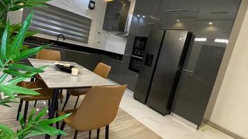Een moderne keuken met donkere kastjes, een ingebouwde oven en een koelkast. Op de voorgrond staat een eettafel met stoelen en een plant.
