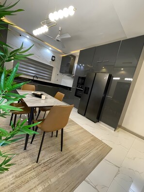 Une cuisine moderne avec des meubles de couleur sombre, un four encastré et un réfrigérateur. On y trouve une table à manger avec des chaises, et une plante est placée au premier plan.
