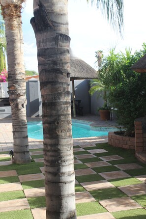 A pool area with palm trees, a thatched-roof structure, and a checkered tile pattern. - Falcon Guesthouse (Upington)