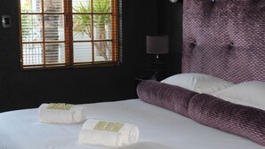 A hotel room with a large bed, two white towels with labels, a window with blinds, a lamp, and a textured wall. - Falcon Guesthouse (Upington)