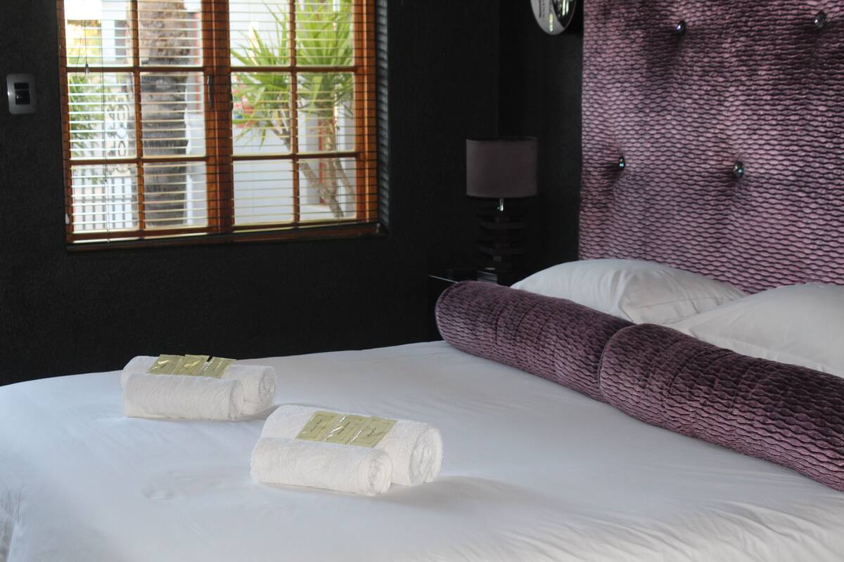 Habitación de hotel con una cama grande, dos toallas blancas con etiquetas, una ventana con persianas, una lámpara y una pared texturizada.