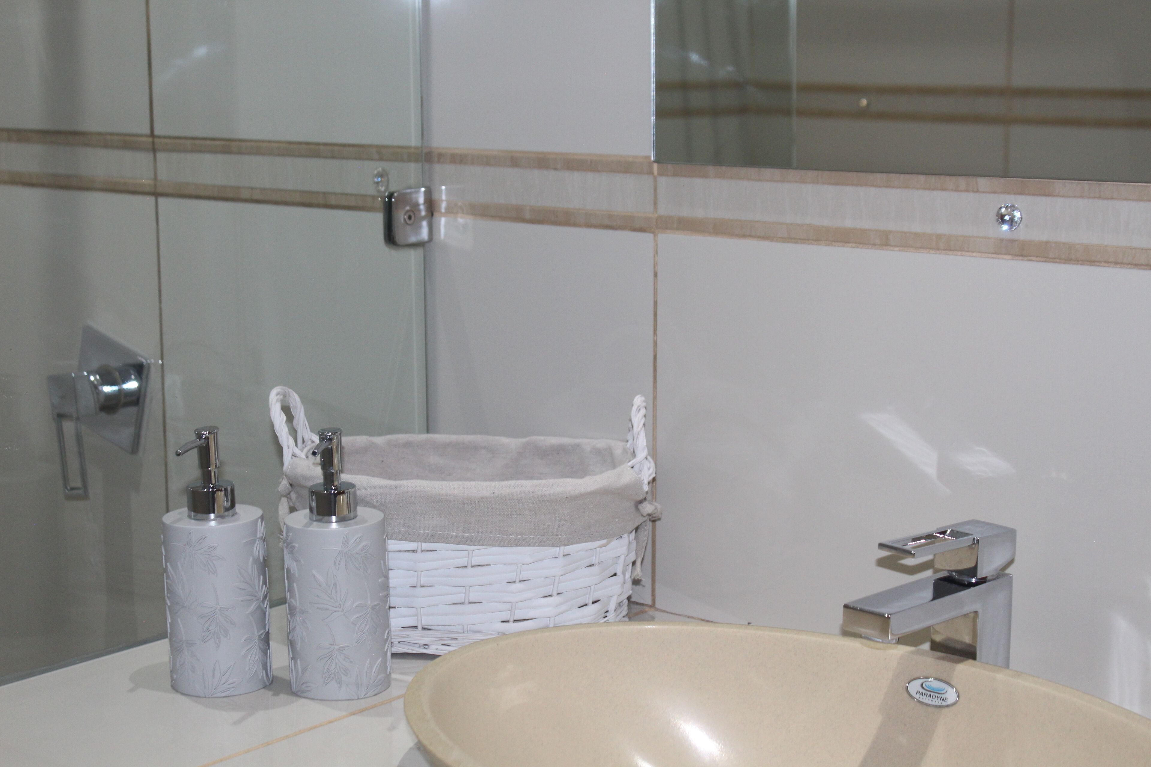 Un baño con lavamanos blanco, dos dispensadores de jabón, una cesta de mimbre blanca y un espejo.