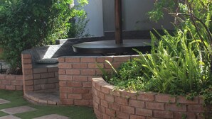 A thatched-roof outdoor seating area with brick walls and green plants. - Falcon Guesthouse (Upington)