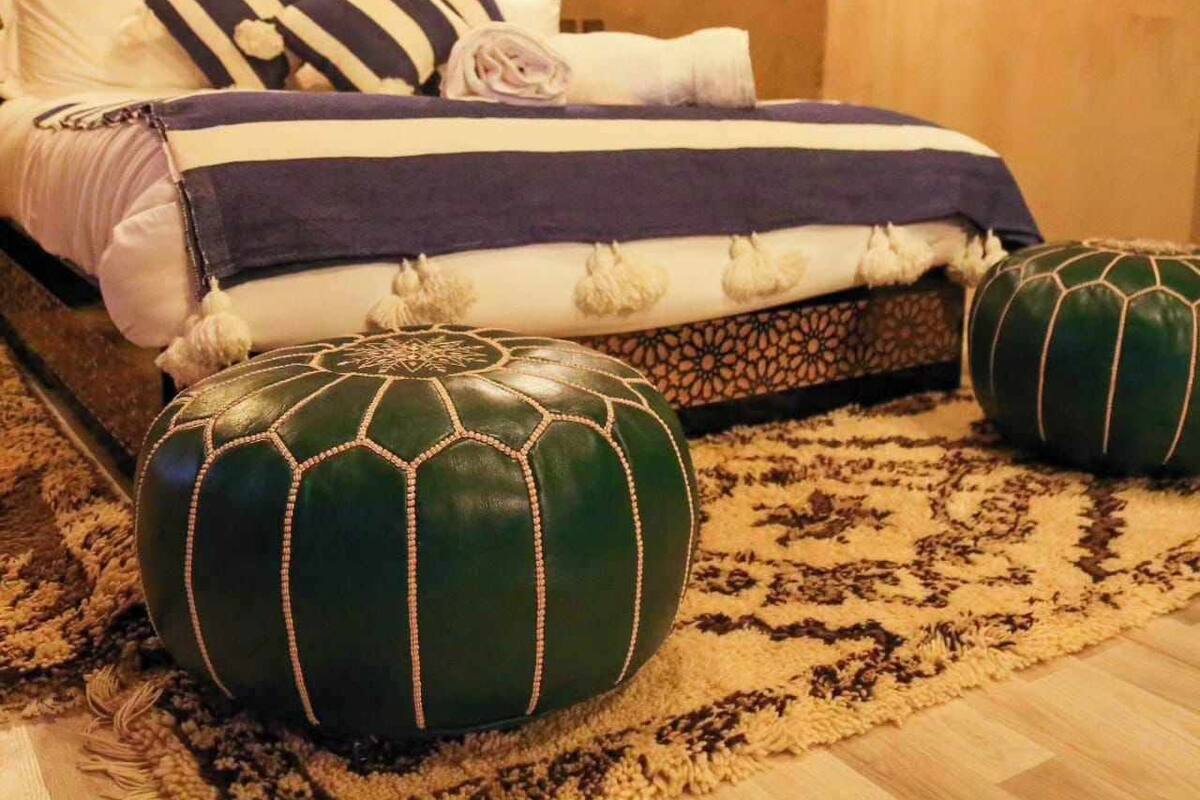 A neatly made bed with striped pillows and a dark blue blanket, two green poufs with tassels, and a patterned rug on a wooden floor.