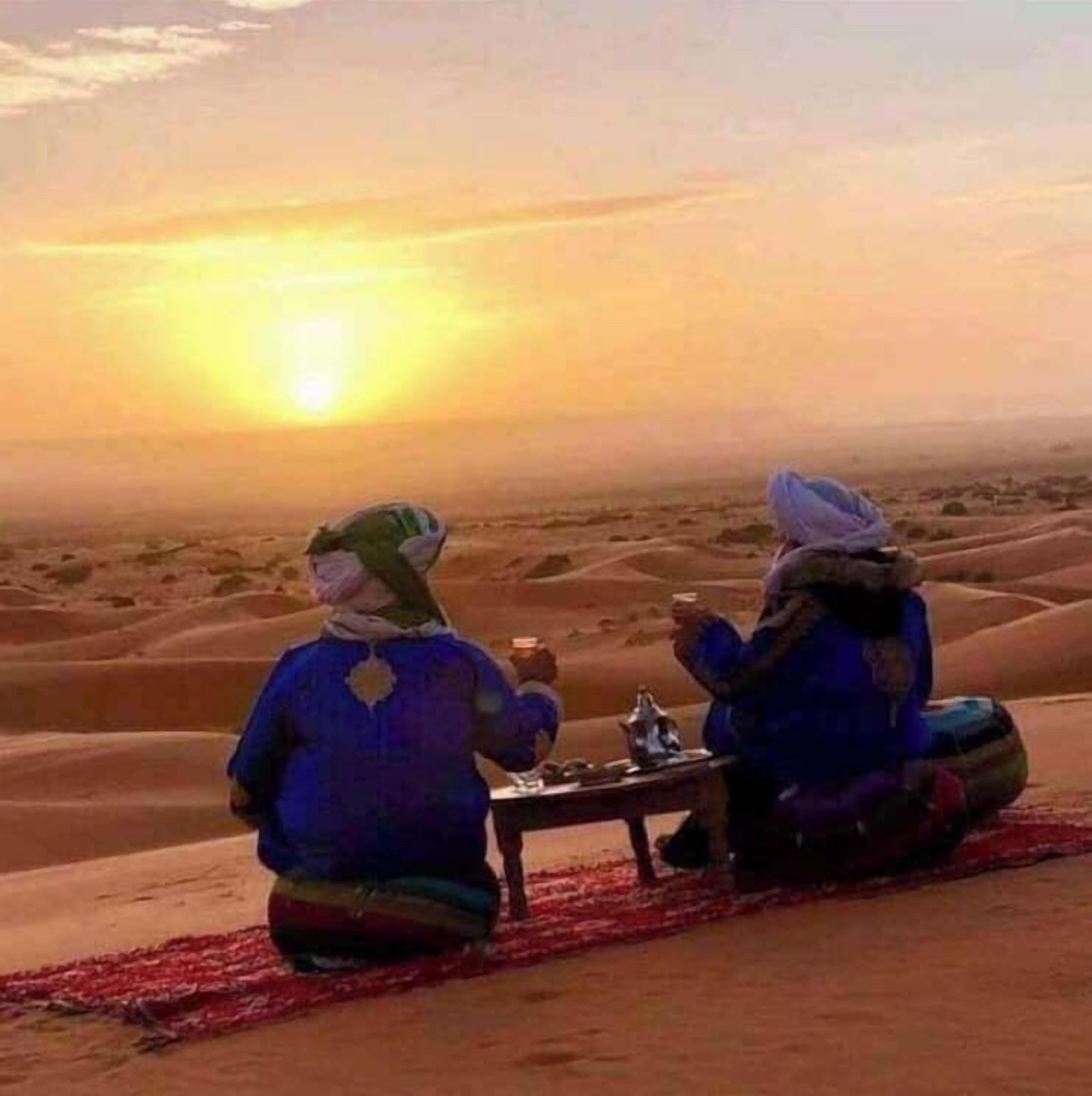 Twee personen zitten op een rood tapijt in een woestijnlandschap met zandduinen, onder een zonsondergang.