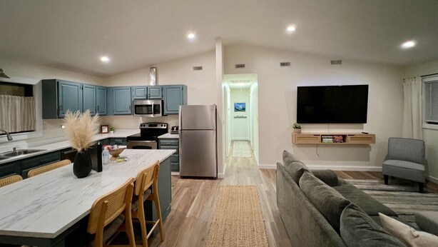 A modern kitchen with a dining area, a living room with a sofa and TV, and a hallway leading to a bathroom. - Wavy Navy - 2 Queen Beds - Centrally Located - Kill Devil Hills OBX (Kill Devil Hills)