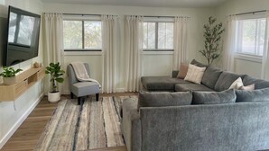 A modern living room with a grey sofa, a flat-screen TV mounted on the wall, a wooden TV stand, a grey armchair, and a large window with white curtains. - Wavy Navy - 2 Queen Beds - Centrally Located - Kill Devil Hills OBX (Kill Devil Hills)