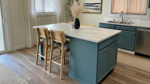 A modern kitchen with a central island, wooden chairs, a vase with dried plants, and a painting on the wall. - Wavy Navy - 2 Queen Beds - Centrally Located - Kill Devil Hills OBX (Kill Devil Hills)