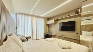 A hotel room with a large bed, a flat-screen TV mounted on the wall, a desk with a chair, and a window with curtains.