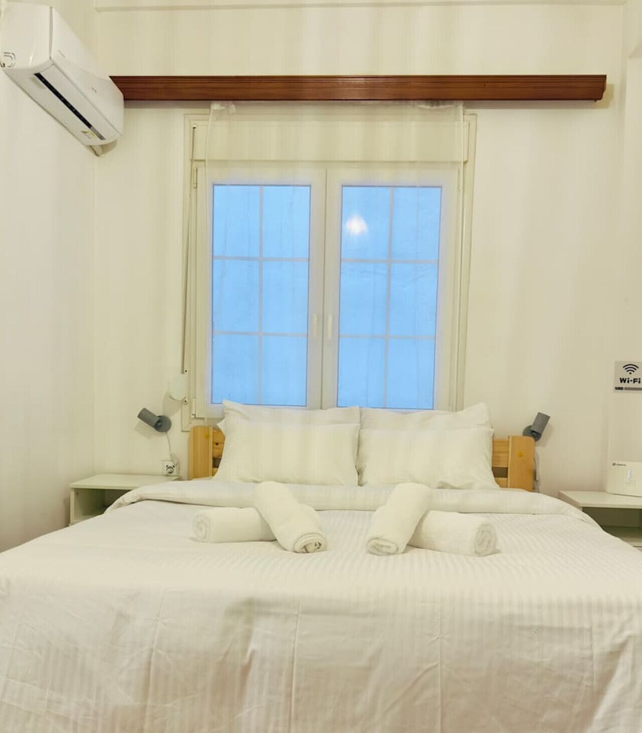 A hotel room with a large bed, two bedside tables, a window with curtains, an air conditioning unit, and a Wi-Fi sign.