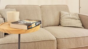 A living room with a beige sofa, a small round wooden table with a book, a white mug, and a colorful abstract painting on the wall.