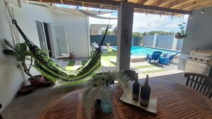 A wooden deck with a hammock, a round table with bottles, and a view of a swimming pool and patio furniture. - Modern 3-bedroom villa with private pool, great alarm system, WiFi, hot water! (Noord)