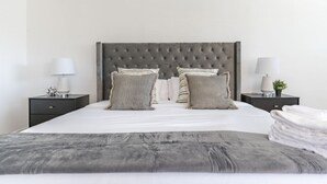 Een netjes opgemaakt bed met een grijs hoofdbord, twee nachtkastjes met lampen, en een grijze plaid over het bed.