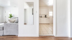 Een moderne woonkamer met een grijze bank, een grote spiegel en een potplant. Een doorgang leidt naar een keuken met witte kasten en een betegelde vloer.