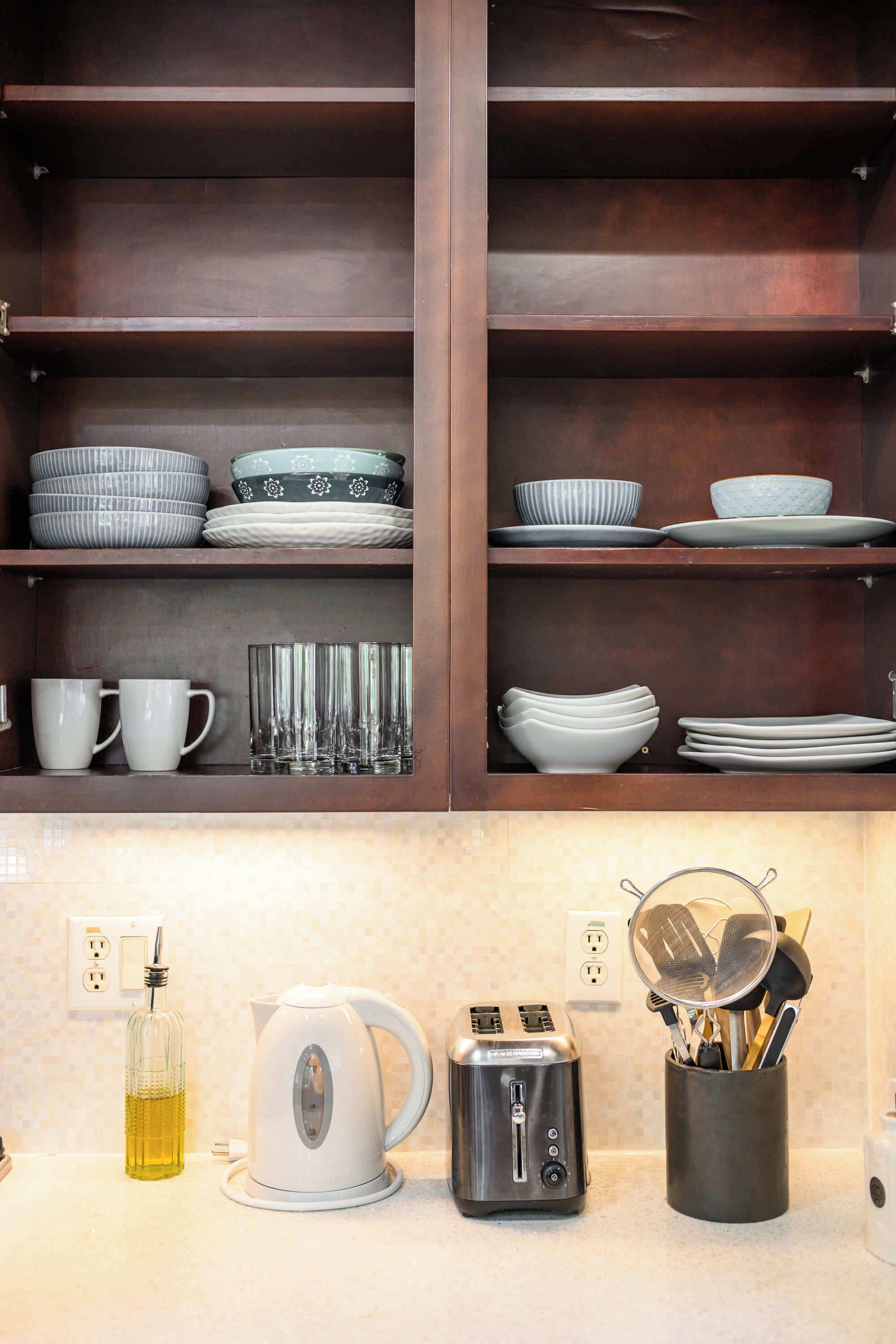 A kitchen with wooden shelves holding various dishes and cups, a kettle, a toaster, and a utensil holder.