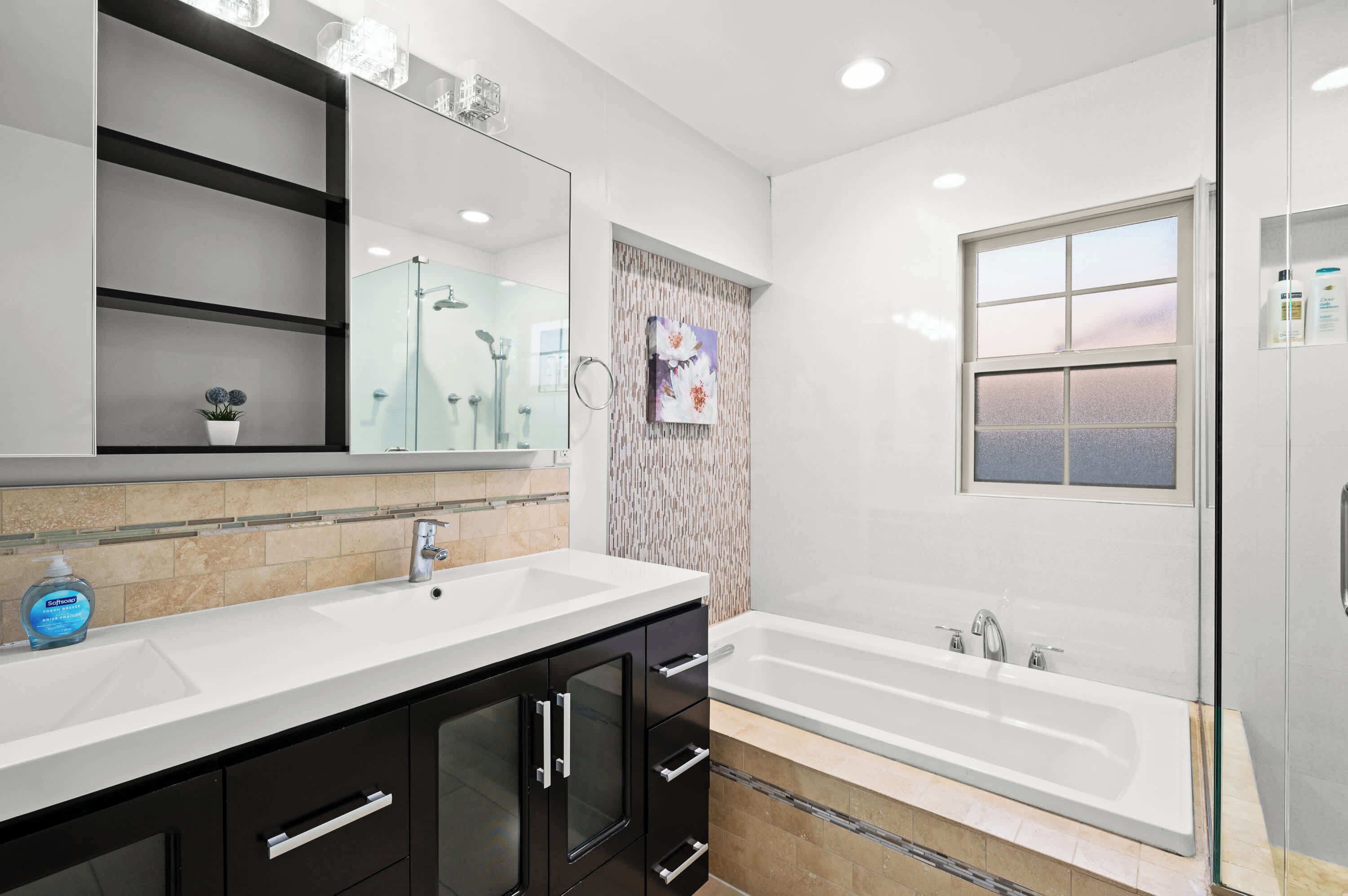 A modern bathroom with a large mirror, a white sink, black cabinets, a glass shower enclosure, and a window with a view.