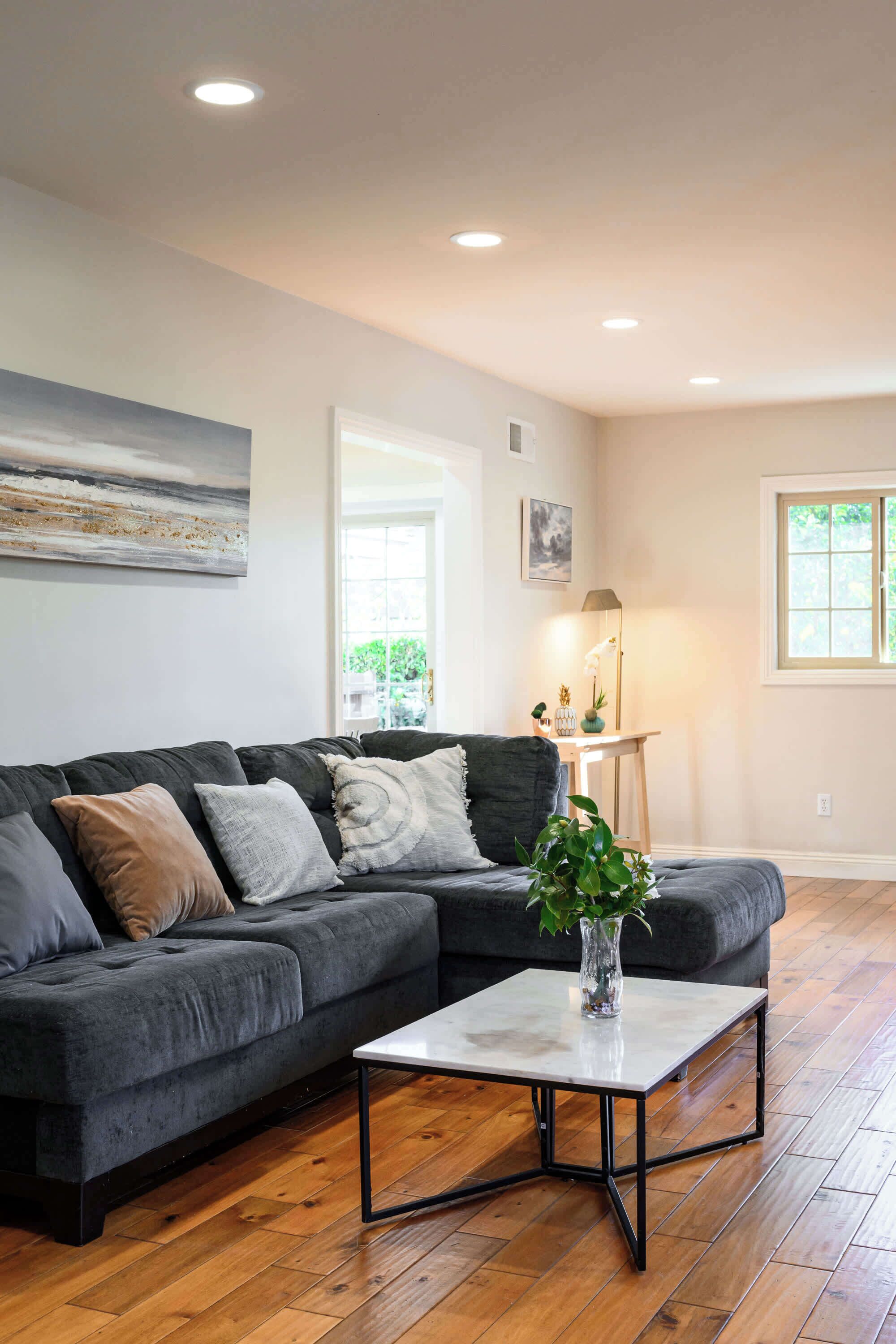 A modern living room with a dark grey sectional sofa, a white coffee table, and a large window with natural light.