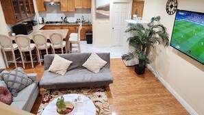 A living room with a grey sofa, a coffee table, and a TV displaying a sports game. There is a kitchen area with wooden cabinets and a dining table with chairs. - Monterey Park Retreat: Spectacular Views & Comfort (Monterey Park)
