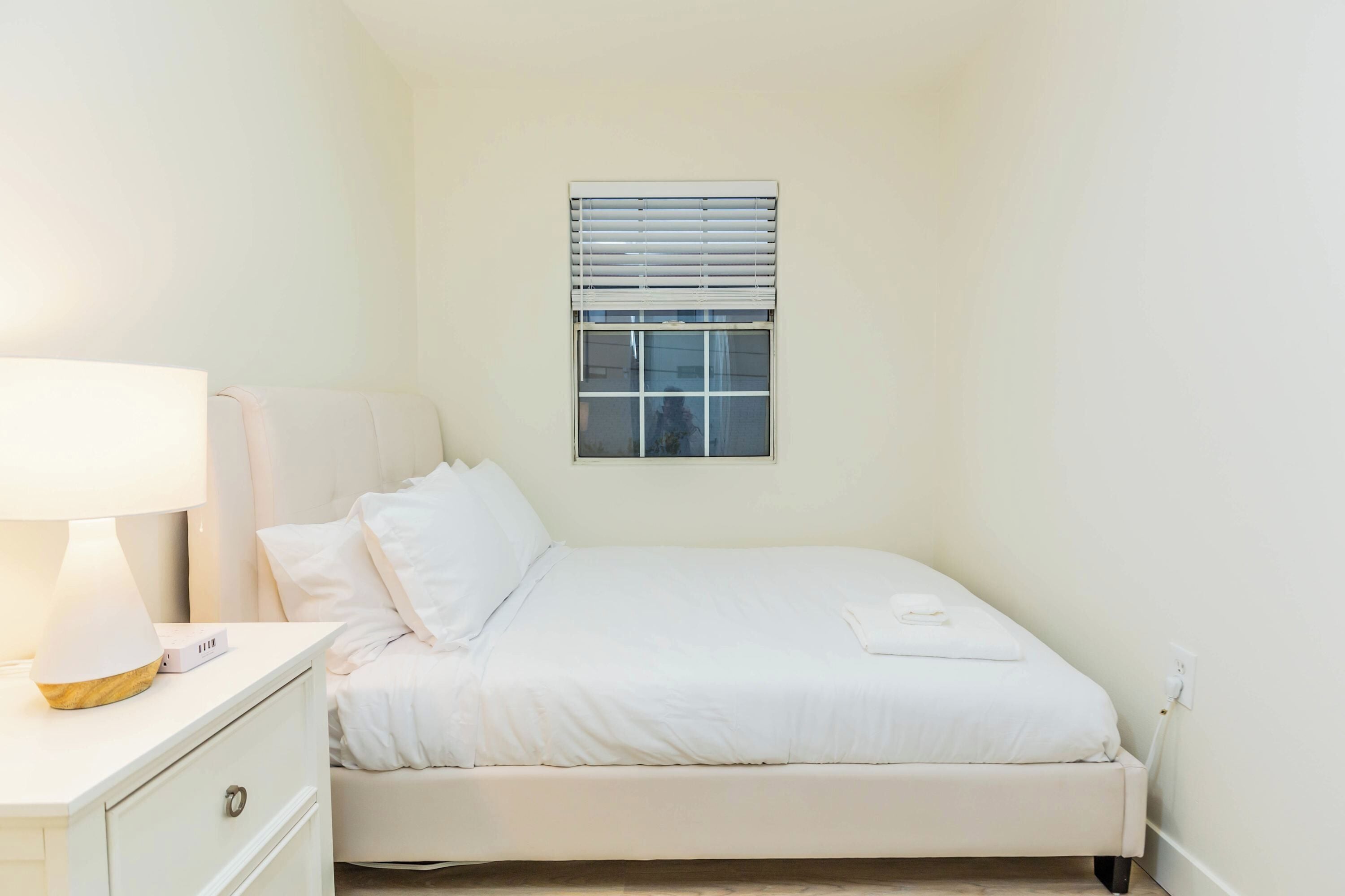 Ein Schlafzimmer mit einem Bett, einem Nachttisch mit Lampe und einem Fenster mit Jalousien.
