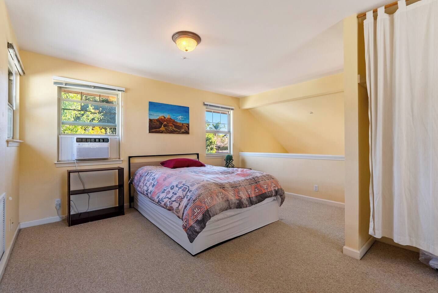 Ein Schlafzimmer mit einem Bett, zwei Fenstern, einer Klimaanlage, einem Bild und einer Deckenleuchte.