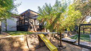 Una casa moderna con fachada de madera, un tobogán amarillo y un patio cercado con árboles.