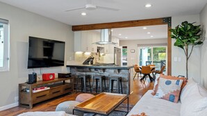 Una sala moderna con televisor, mesa de centro de madera y un sofá con cojines estampados. Se observa un área de cocina con taburetes de bar al fondo.