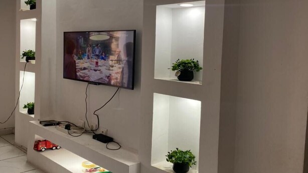 A modern living room with a flat-screen TV mounted on a white wall, surrounded by recessed lighting and small shelves with potted plants. - Douala_yassa_meublé (Douala)