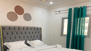 A neatly made bed with a grey upholstered headboard, white bedding, and a black furry rug at the foot of the bed. - Douala_yassa_meublé (Douala)