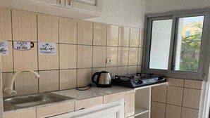 A small kitchen with light wooden cabinets, a stainless steel sink, a kettle, and a window providing natural light. - Douala_yassa_meublé (Douala)