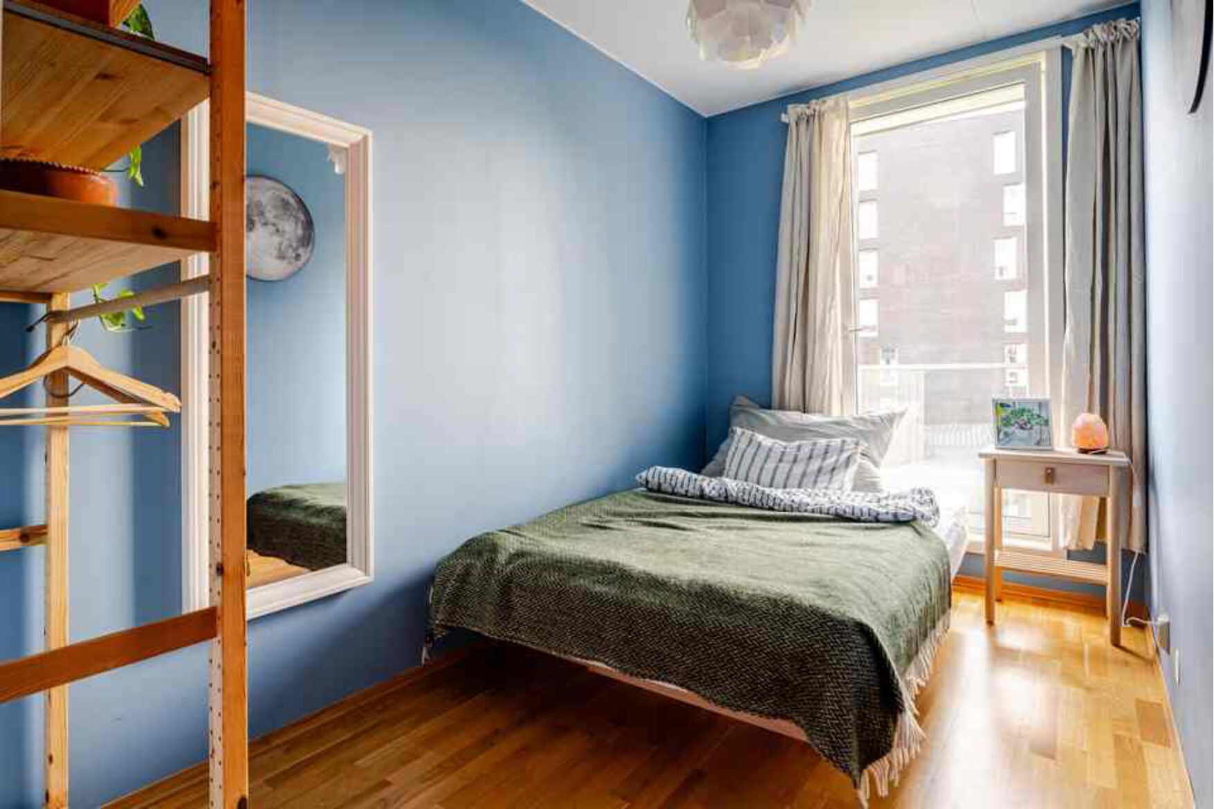 A bedroom with a bed, a wooden shelf, a mirror, and a window with curtains.