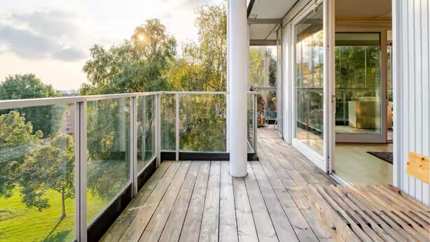 A modern balcony with glass railings, wooden flooring, and a view of trees and buildings. - Spacious 3-bedroom with 3 balconies, 2 bthrms, sea view and free indoor parking (Oslo)
