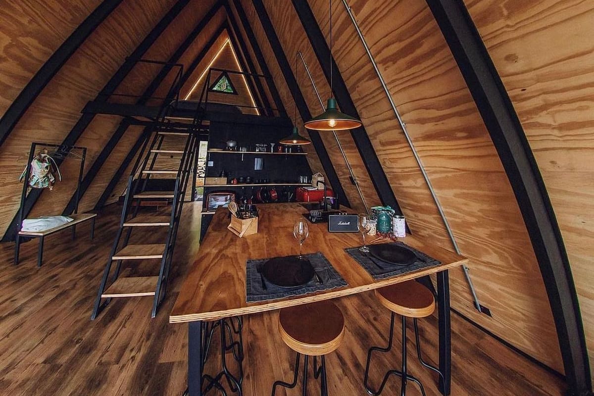 A cozy attic-style room with a wooden interior, a dining table with chairs, a ladder, and a hanging light fixture.