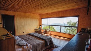 Un dormitorio con un ventanal grande, paredes de madera, una cama, una silla y un escritorio con una lámpara.