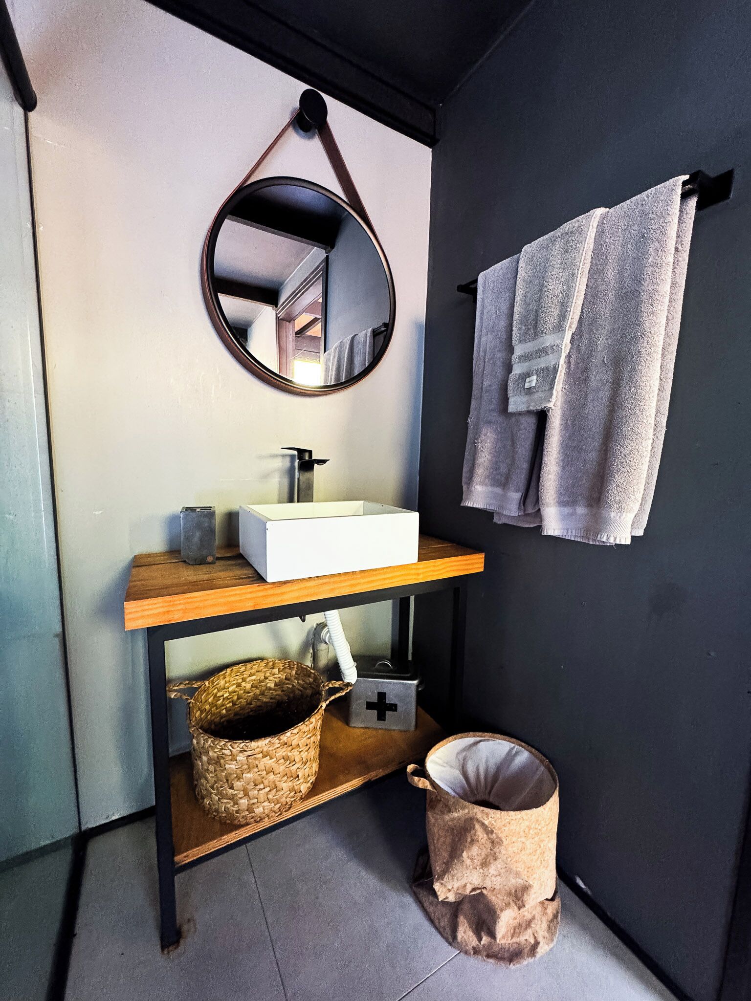 Une salle de bain avec un lavabo blanc, un miroir rond, un porte-serviettes avec deux serviettes, un panier en osier et une poubelle.